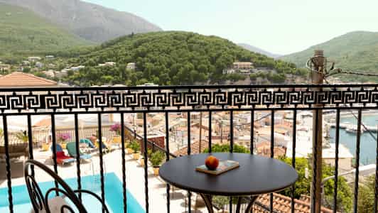 Utsikt över en balkong med ett bord och dekorativt räcke, poolområde, hustak och grönskande kullar. (AI-genererad text)