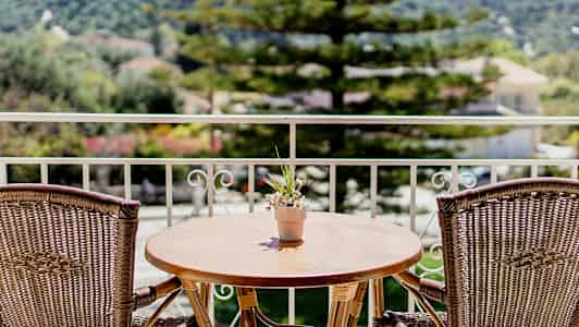 En balkong med ett träbord och två rottingstolar, och en liten blomkruka med blommor på bordet. I bakgrunden syns träd och byggnader. (AI-genererad text)