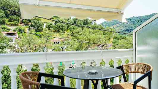 Utsikt från en balkong med ett litet runt bord och två stolar, omgiven av grönska och ett kuperat landskap. (AI-genererad text)