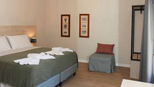 Ett hotellrum med en säng, två tavlor på väggen, en stol med kudde och en öppen garderob. (AI-genererad text)