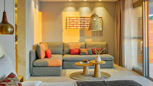 Ett vardagsrum med en grå hörnsoffa, rödaktiga kuddar, ett par små träbord och en dekorativ matta. En hängande lampa och en målning på väggen syns också. (AI-genererad text)