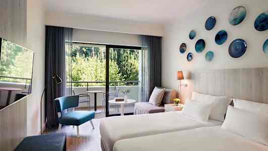 Modernt inrett hotellrum med två sängar, en sittgrupp och balkong med utsikt över grönska. (AI-genererad text)