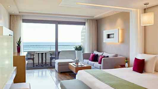 Ett hotellrum med modern inredning, som inkluderar en säng, sittgrupp och en utsikt över havet genom glasdörrar. (AI-genererad text)