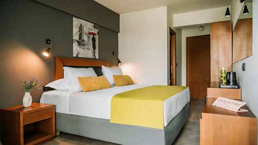 Ett sovrum med en dubbelsäng med vita och gula sängkläder, matchande kuddar, en nattduksbord med en vas av blommor, och en vägghängd tavla ovanför sängen. (AI-genererad text)