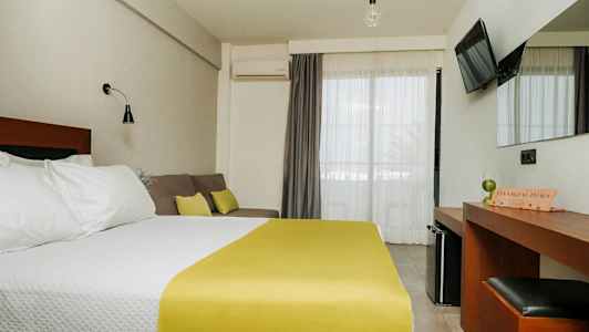 Ett hotellrum med en dubbelsäng med vita sängkläder och gult överkast, en soffa med kuddar, ett skrivbord med spegel, och en TV på väggen. Balkongdörrar med ljusa gardiner. (AI-genererad text)