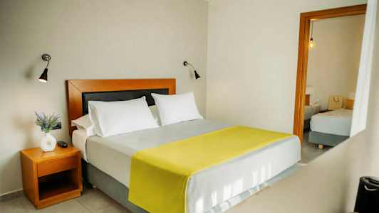 Sovrum med en dubbelsäng, två vita kuddar och en gul filt. Ett sidobord med en vit vas och en växt. (AI-genererad text)