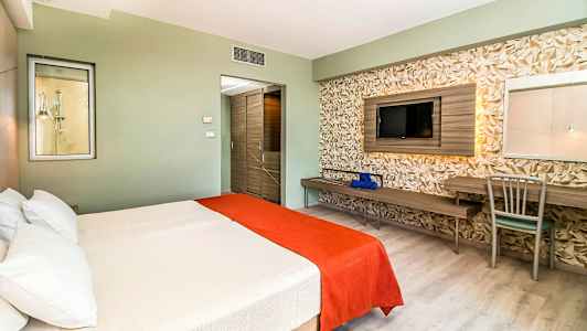 Hotellrum med en dubbelsäng täckt med vit och orange sängkläder, en vägg med geometrisk dekor, samt en tv och skrivbord. (AI-genererad text)