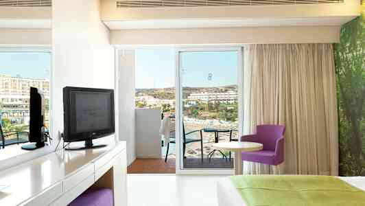 Hotellrum med utsikt mot havet, möbler inkluderar en TV, ett skrivbord, en lila stol och en stor säng. (AI-genererad text)