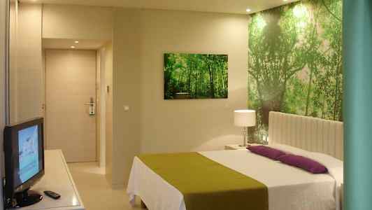 Ett hotellrum med en säng, grön och vit inredning, en TV och en väggdekoration föreställande en skog. (AI-genererad text)