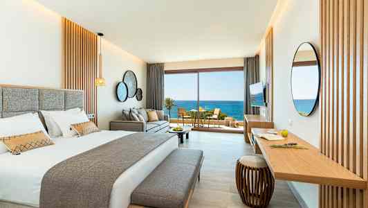 Ett stilrent hotellrum med en dubbelsäng, soffa och ett skrivbord. Stora glasdörrar öppnar upp till en balkong med utsikt över havet. (AI-genererad text)