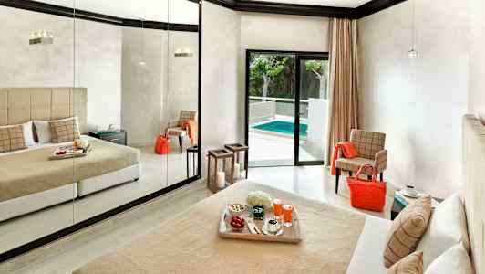 Ett hotellrum med en stor spegelvägg, en säng med beige överkast och kuddar, och en balkong dörr som leder ut till en pool. (AI-genererad text)