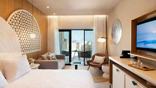 Ett modernt hotellrum med ljusa möbler och dekoration, inkluderande en säng, soffa, stol och TV. Rummet har stora glasdörrar som leder ut till en balkong. (AI-genererad text)