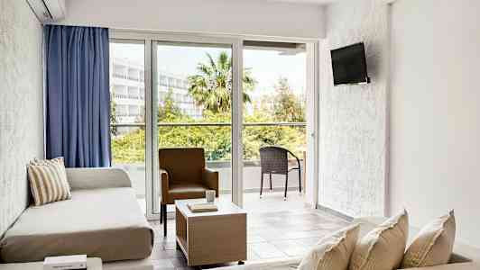 Ett ljust hotellrum med sittgrupp, soffor, en stol, ett soffbord och stora fönster som vetter ut mot en balkong med utemöbler i en grönskande miljö. (AI-genererad text)