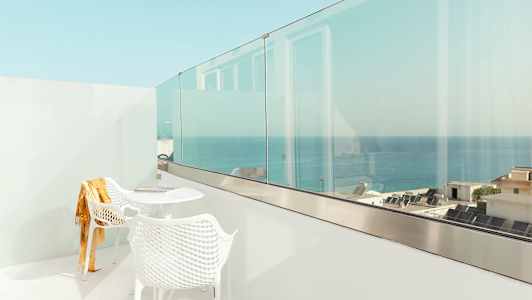 En vit balkong med två stolar och ett bord, med utsikt över havet och byggnader. (AI-genererad text)