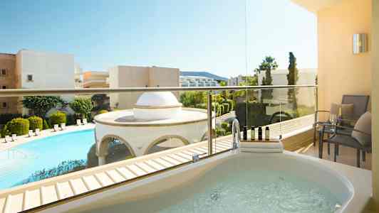 Bild av en balkong med ett spabadkar och sittmöbler. Utsikt över en pool och omgivande byggnader. (AI-genererad text)