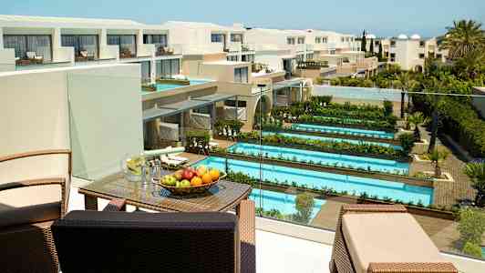 Hotellområde med flera pooler och grönområden, samt en terrass med möbler och frukt. Utsikt över en modern byggnad. (AI-genererad text)