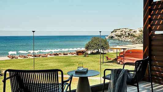 Utsikt från en terrass över grönt gräs, en strand med solstolar och parasoller samt havet och klippor i bakgrunden. (AI-genererad text)