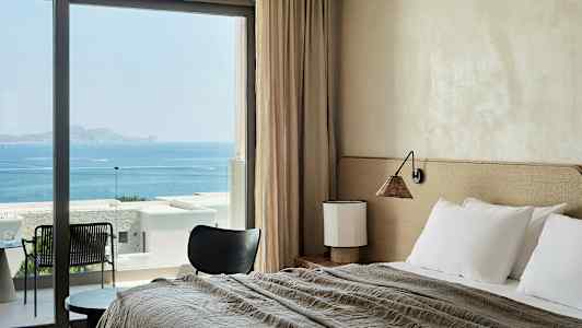 Ett hotellrum med säng, bord och stol, med havsutsikt genom stora fönster. (AI-genererad text)