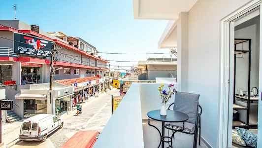 En balkong med bord, stol och blommor i förgrunden, samt en vy över en gata med butiker och byggnader. (AI-genererad text)