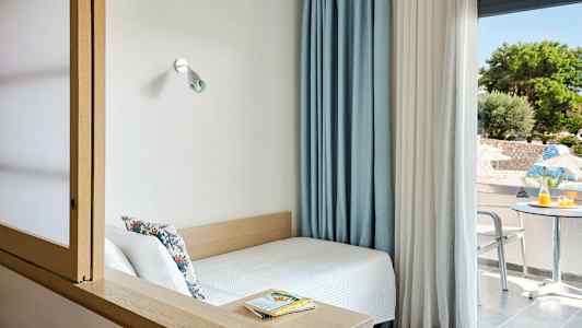 Ett litet rum med en säng vid ett fönster och utsikt över en uteplats med bord och stolar. (AI-genererad text)