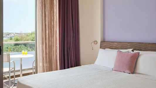 Ett hotellrum med en bäddad säng, lila vägg och gardiner samt en balkong med bord och två stolar utsikt mot grönska. (AI-genererad text)