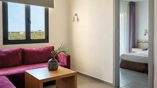 Ett rum med lila soffhörn, ett bord med en vas och ett angränsande sovrum synligt genom en dörröppning. (AI-genererad text)