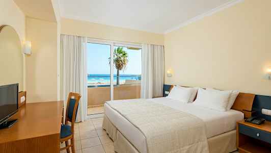 Hotellrum med en dubbelsäng, skrivbord, stol och utsikt över en strand genom en glasdörr. (AI-genererad text)