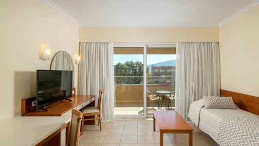 Ett hotellrum med en TV på ett skrivbord, två enkelsängar, och en balkong med utemöbler. (AI-genererad text)