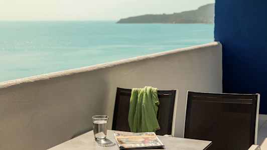 Två stolar med en grön filt på den ena, stående vid ett bord som har ett glas vatten och en tidning, med havsutsikt. (AI-genererad text)