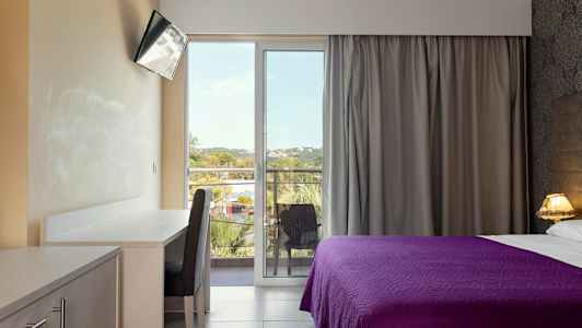 Ett hotellrum med ett skrivbord, en stol, en TV och en säng med lila överkast. Rummet har en balkong med utsikt. (AI-genererad text)