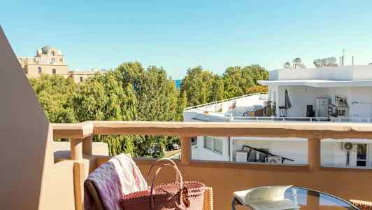 Utsikt från en balkong mot träd och byggnader. Ett bord med en bok och en stol med en väska i förgrunden. (AI-genererad text)