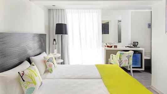 Ett ljust hotellrum med två enkelsängar, dekorativa kuddar och en limegrön sängfilt, samt en sittplats och ett skrivbord med spegel. (AI-genererad text)