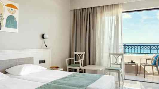 Hotellrum med dubbelsäng, två stolar och ett bord vid fönster med havsutsikt. (AI-genererad text)