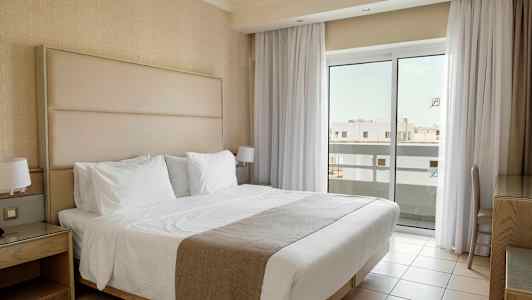 Ett hotellrum med en dubbelsäng klädd i vita sängkläder, nattduksbord och stora fönster med gardiner som släpper in ljus. (AI-genererad text)