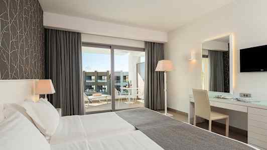 Ett hotellrum med två enkelsängar, skrivbord, stol och en fönstervy mot en balkong med bord och stolar. (AI-genererad text)