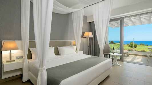 Hotellrum med en bred säng, vita gardiner, och utsikt mot havet genom stora fönster. (AI-genererad text)