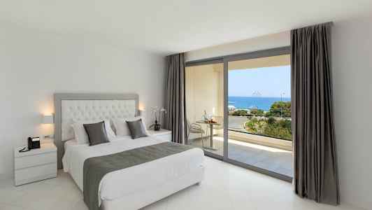 Ett sovrum med en dubbelsäng och utsikt över havet genom stora fönster. (AI-genererad text)