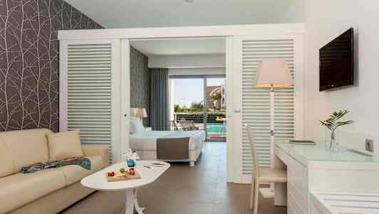 Ett hotellrum med soffa, bord med dekorativa föremål, skrivbord och stol. Sängen i bakgrunden leder till en terrass med utsikt över en pool. (AI-genererad text)