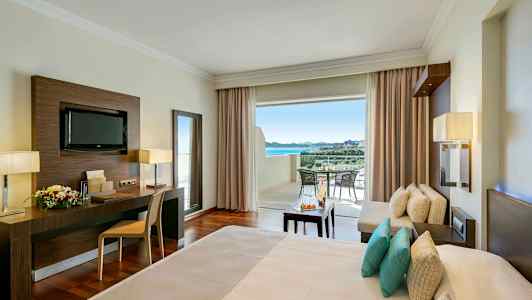 Ett hotellrum med en bäddad säng, arbetsområde, sittgrupp och öppen balkong med utsikt. (AI-genererad text)