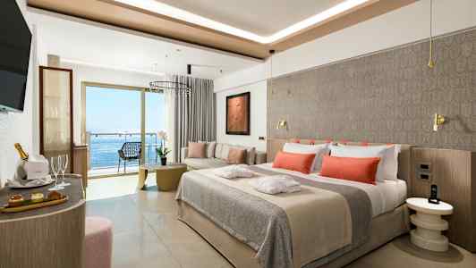 En modern hotellsvit med en bäddad säng med orange kuddar, sittgrupp och balkong med havsutsikt. (AI-genererad text)