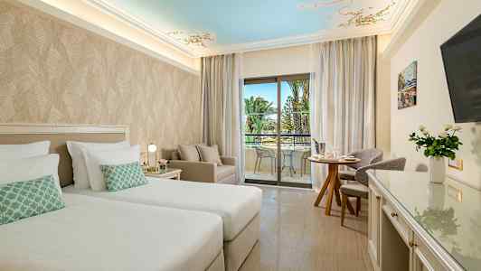 Ett hotellrum med två sängar, sittgrupp och balkong med utsikt mot palmer. (AI-genererad text)