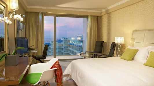 Hotellrum med en dubbelbädd säng, stolar och utsikt över balkonger och havet genom stora fönster. (AI-genererad text)