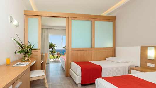 Ett hotellrum med två enkelsängar, en arbetsyta med en stol och en glasvas med blommor. Rummet har en skjutdörr som leder till en balkong med utsikt över havet. (AI-genererad text)