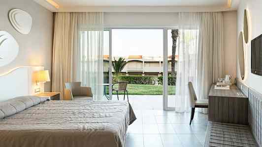 Ett hotellrum med säng, skrivbord, stol och en balkongdörr som vetter mot en trädgård. (AI-genererad text)