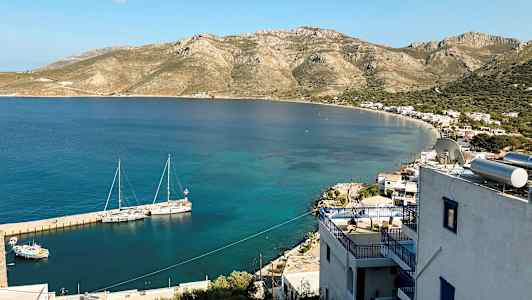 Vy över en kust med små byggnader och berg i bakgrunden. Båtar ligger förtöjda vid bryggan i det klara vattnet. (AI-genererad text)