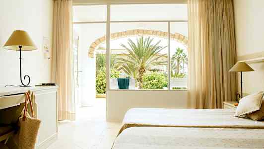 Ett hotellrum med två sängar, skrivbord och lampa. Fönster och dörr leder till en trädgård med palmer. (AI-genererad text)