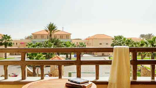 Utsikt från en balkong med ett bord och handduk över balkongräcket, omgivande byggnader och palmer syns i bakgrunden. (AI-genererad text)