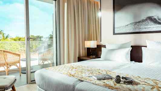 Ett hotellrum med en dubbelsäng, en glasdörr som leder till en balkong med stolar. (AI-genererad text)