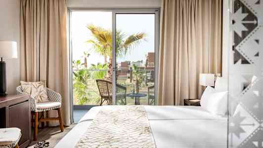 Ett hotellrum med en stor säng, en sittgrupp och en balkong med utsikt över palmträd. (AI-genererad text)