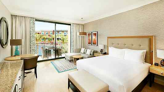 Ett hotellrum med en stor säng, sittgrupp och balkong med utsikt över en pool och byggnader. (AI-genererad text)